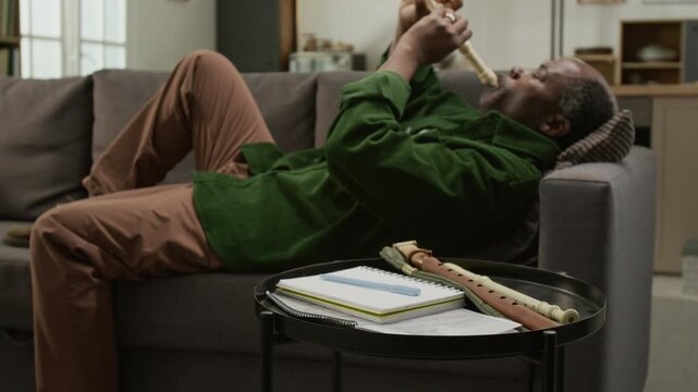 Side view shot of senior African American man playing recorder while relaxing on couch with focus on music notes on coffee table in foreground, copy space Suitable for lifestyle product advertising