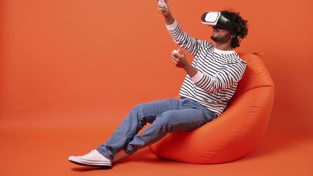 Young amazed Indian man he wears striped sweatshirt casual clothes sit in bag chair watching in vr headset pc gadget isolated on plain orange red color background studio portrait. Lifestyle concept