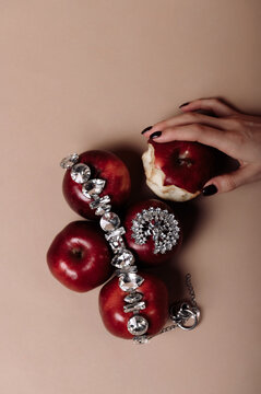 Close-up overhead view of a woman's hand holding a half eaten red apple next to a stack of apples and jewellery
