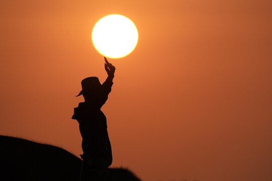 Side view silhouette of a mahout riding an elephant at sunset with his arms raised pretending to warm his hands on the sun, Thailand