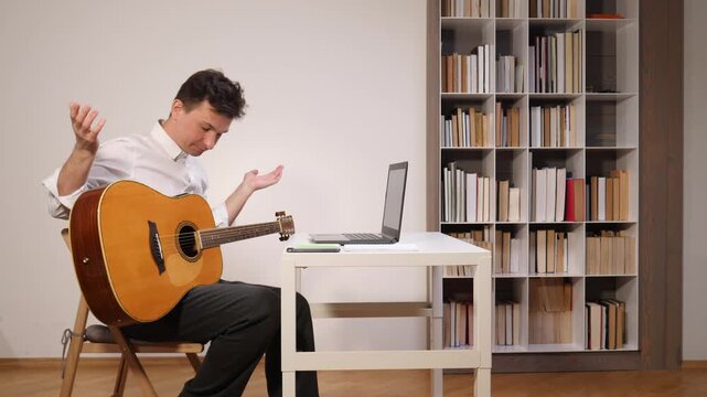 Man holding an acoustic guitar looking frustrated and confused while taking an online music lesson on his laptop, sitting at a desk with a bookshelf filled with books in the background