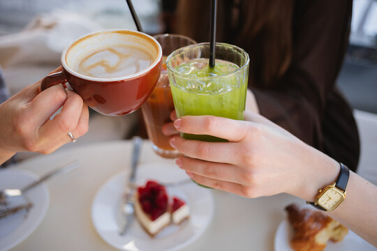 Close-up of three women sitting at a table clinking glasses of iced matcha latte, espresso tonic and a cappuccino