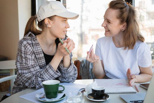 Two smiling young women sitting in a cafe with a laptop and assorted paperwork brainstorming a project