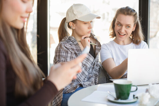 Three young women working in a cafe with two of them looking at a laptop computer while another is using her mobile phone
