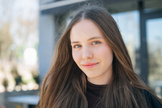 Close-up portrait of a smiling young woman with long brown hair standing in front of a building on a sunny day