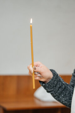Close-up of a woman's hand holding a single lit yellow beeswax candle during a religious service or prayer. Minimalist composition with a soft blurred background, symbolizing faith, hope, and spiritua