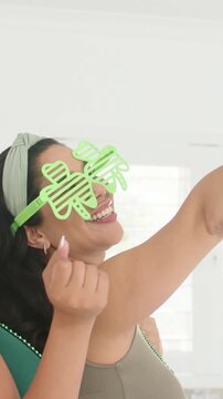 Vertical video: Smiling woman in green tank shamrock specs taking selfie in kitchen as cell appears
