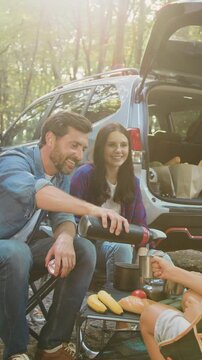 Father mother and children bonding in forest nature, drinking hot tea witch thermos sitting on table near parking car in sunny day. Attractive bearded man pouring hot drink from thermos to pretty kids