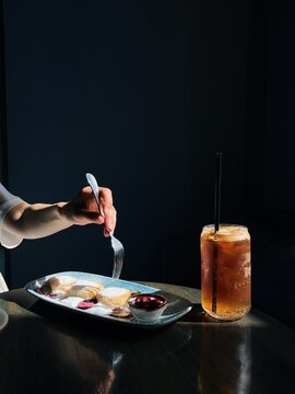 Side view of a woman sitting at a table eating mini Syrniki pancakes with yogurt and blueberries served with a refreshing iced espresso tonic