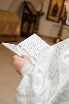 Close-up of an Orthodox priest's hands holding an open prayer book with Ukrainian text. The priest is wearing a traditional white embroidered liturgical vestment. Soft focused church interior in the b