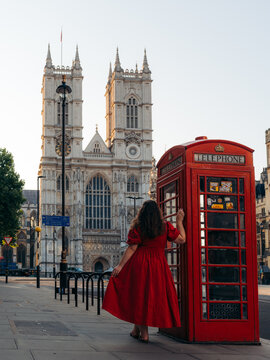 View of a woman in a vibrant red dress pauses by a classic red telephone box, with the majestic Westminster Abbey rising in the background, London, England, United Kingdom.