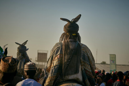 Zaria City, Nigeria - 22 March 2026: View of a prominent figure in elaborate traditional attire with distinctive horned headwear, amidst a bustling crowd under a soft, clear sky.