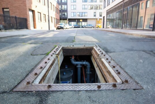 Open manhole with pipes and barrels in an urban alleyway with modern buildings