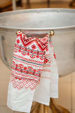 A white towel with red and black geometric embroidery (rushnyk) hanging on the handle of a large metal baptismal font. A symbol of Ukrainian national traditions during the Orthodox sacrament of baptis