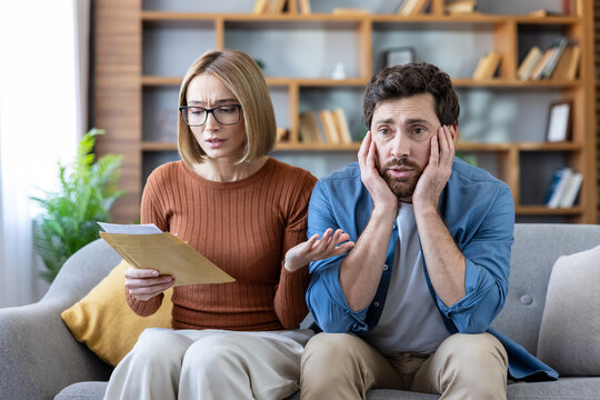 Young couple sitting on a sofa at home, feeling stressed and anxious after opening an important letter containing bad news about finances, bills, or debt