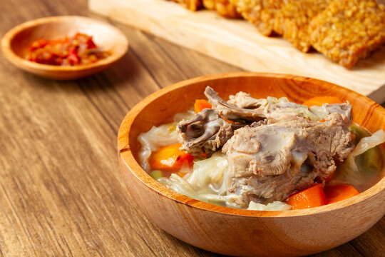 Warm wooden bowl holds rich, savory stew. Crispy Tempeh sit beside the bowl. sambal condiment. All rests on rustic wooden table. Cozy, hearty meal suggests comfort. traditional indonesian food