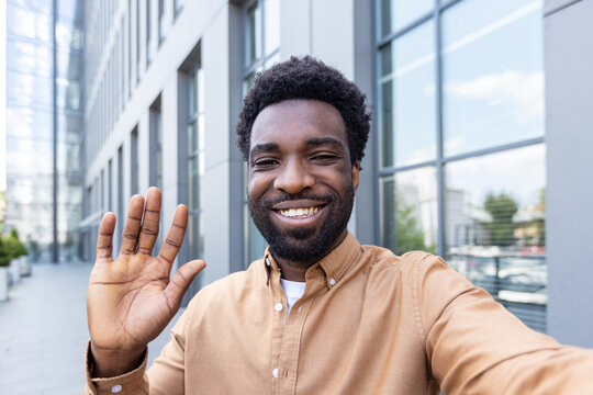 Smiling man with a groomed beard and textured hair happily waving a hand, taking a selfie or video calling outdoors in front of a modern corporate building, representing communication and connection