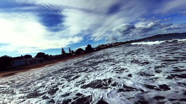 Aerial FPV view of the dramatic dark waves crashing onto the reddish-brown sand, with distant town buildings under a vibrant blue sky, Galatas, Chania, Greece.