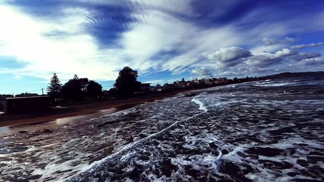 Aerial FPV view of the dynamic coastline, where deep blue waves crash against the golden sand beach, with white buildings under a vibrant sky, Galatas, Chania, Greece.