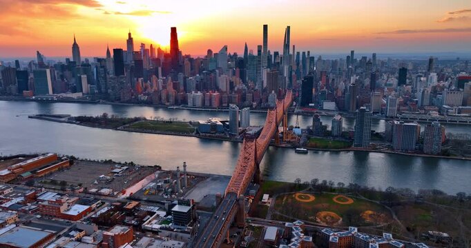 Approaching the gorgeous Queensboro Bridge over the calm East River. Stunning skyline of Upper East Side neighborhoods in Manhattan against orange sky.