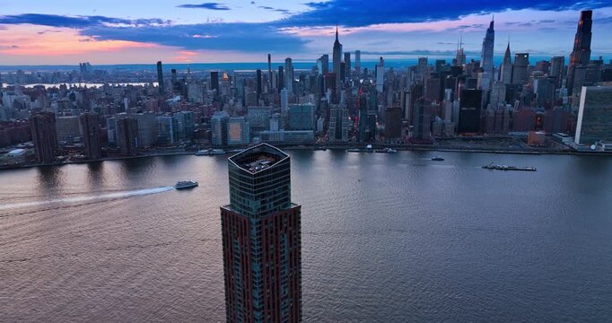 Descend along the Gotham Point skyscraper in Queens, New York, USA. Boats move by the riverscape along Manhattan skyline at dusk time.
