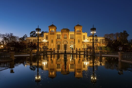 Illuminated art museum Museo de Artes y Costumbres Populares de Sevilla reflected in a fountain, blue hour, Plaza de America, Seville, Andalusia, Spain