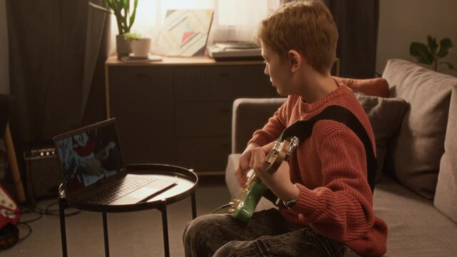 Warm toned side view shot of child playing electric guitar while watching music videos on laptop screen pat home, young child prodigy, copy space Useful for talent development programs