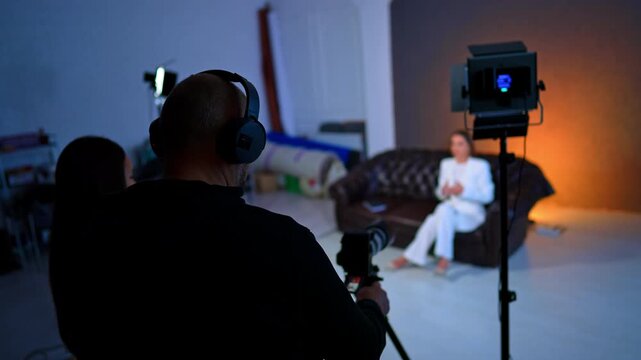 Rear view of the cameraman taking video in the studio. Woman in white suit sits on the sofa talking to camera. Blurred backdrop.
