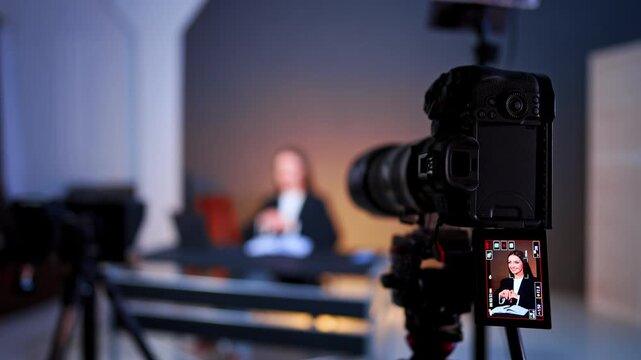 Professional camera taking video of a beautiful woman sitting at desk. Blurred backdrop. Blog footage in photo studio.