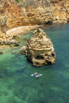 Two SUPs in the turquoise sea, cliffs of sandstone rocks, rock formations and sandy beach, Praia da Marinha, Algarve, Lagos, Portugal