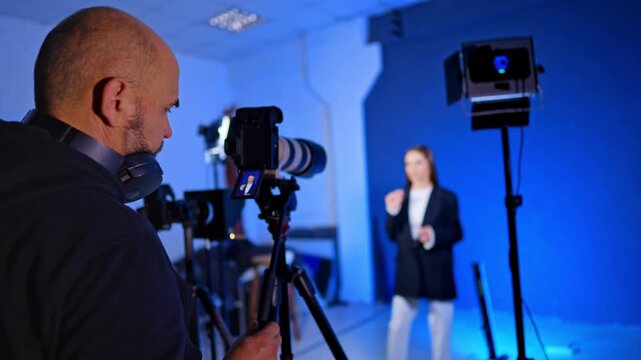 Caucasian man in headphones stands near camera. Cameraman is recording a video with a woman talking in the blurred backdrop against blue wall.
