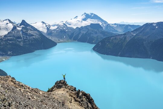 Young man standing on a rock, stretching his arms in the air, view of mountains and glacier with turquoise blue lake Garibaldi Lake, peaks Panorama Ridge, Guard Mountain and Deception Peak, Garibaldi Provincial Park, British Columbia, Canada