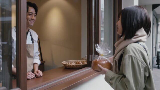Customer reaching to buy loaf, smiling baker in apron handing wrapped round loaf at bakery window