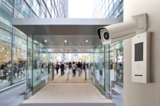 A surveillance camera is positioned outside a large glass entrance where many people walk by. It is a sunny day in a busy urban area with tall buildings