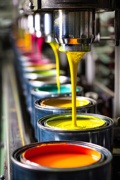 Paint is being poured into cans on a factory production line. The equipment is clean and shiny, highlighting the paint colors being collected