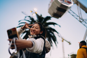 Obraz premium Happy Asian woman posing for selfie at carnival, victory sign, mobile blogging, 5G connection, authentic representation, emotional joy, inclusion, digital lifestyle, technology, real people, fun