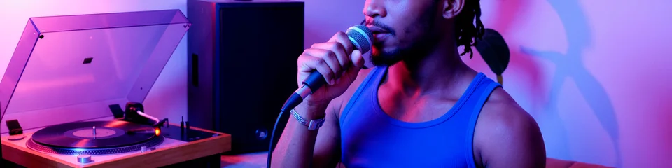 Close up of young Black man singing into microphone beside turntable, recording music content in studio. Ideal for audio production, nightlife promotion, entertainment marketing © Mediaphotos