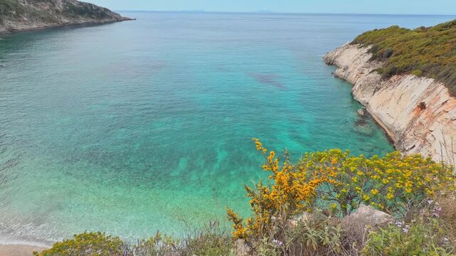 Turquoise Ionian waters at Filikuri Beach, Albania. Scenic coastal view featuring yellow spring wildflowers, rugged cliffs, and the crystal-clear sea with empty beach of Albanian Riviera.
