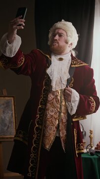 Vertical medium long shot of young bearded Caucasian man in wig and embroidered baroque coat trying to take selfies on smartphone in classical studio