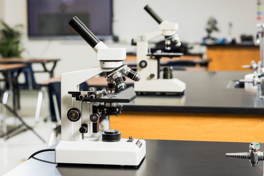 Row of white compound microscopes on black lab benches in an empty high school science classroom ready for student use