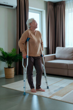 Vertical shot of senior Asian woman standing with crutches at home, recovering from leg injury and practicing mobility. Useful for rehabilitation, home care, recovery support marketing