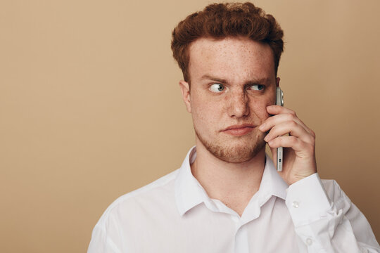 Young man with red hair and freckles wearing white shirt talks on smartphone with serious expression. Neutral beige background. Concept of communication, technology, and thoughtful mood.