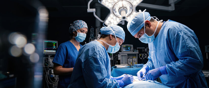Two surgeons and an assistant work together in an operating room. They wear masks and gloves while focusing on a surgical task. Medical equipment surrounds them