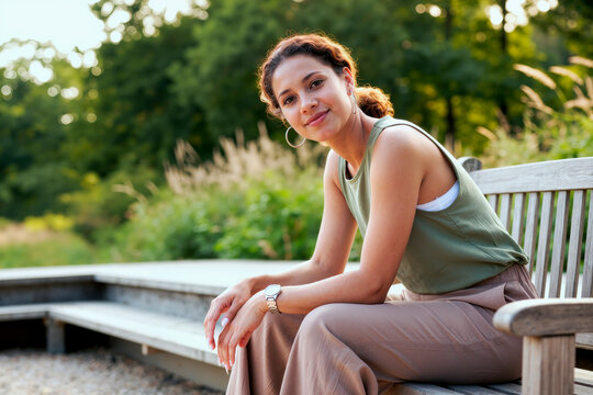 Portrait of young woman sitting on park bench, looking at camera and relaxing outdoors, conveying wellbeing and balance. Useful for mental health, self care, lifestyle campaigns