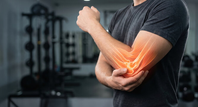 A man stands in a gym and holds his elbow with one hand. He appears focused and seems to be experiencing discomfort in his joint. The setting shows fitness equipment
