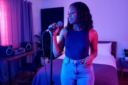 Young Black woman singing into microphone in home recording setup, creating music content for digital audience. Ideal for creator economy, streaming, podcast promotion, vertical