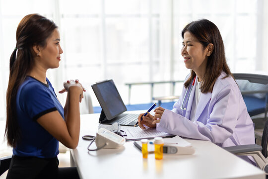 Asian female doctor explaining diagnosis to her patient during a medical consultation in a clinic, using a tablet and taking notes on a document.