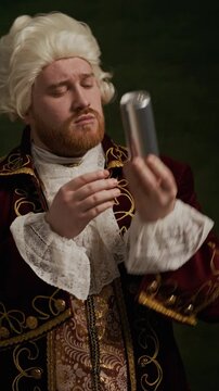 Vertical shot of young Caucasian aristocrat wearing embroidered rococo coat and wig holding and examining drink can indoors