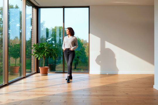 Wide shot of young woman standing near floor to ceiling windows in empty home, considering property move or renovation. Ideal for real estate, relocation, interior marketing