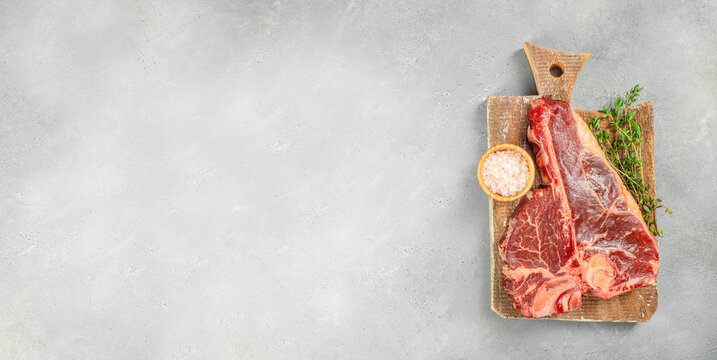 Raw porterhouse t-bone steak on wooden cutting board with salt and herbs on light concrete background with copy space